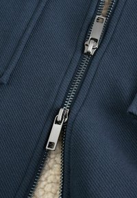 Námornícky modrý zipsový odev s textúrovanou tkaninou, obsahujúci metalický zips a mäkkú fleecovú podšívku. Detaily ukazujú zuby zipsu a ťahák.