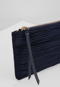 Pochette bleu marine en tissu plissé texturé, équipée d'une fermeture éclair dorée et d'une fine lanière en cuir pour poignet. Finition lisse, forme rectangulaire.