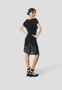 Melna ceļa garuma kleita ar īsām piedurknēm, rotāta ar smalkām pelēkām un sarkanām ziedu rakstiem apakšmalā. Valda ar melnām ķīļveida sandales.