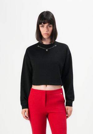 Woman with straight dark hair wearing a black cropped sweatshirt with chain detail and high-waisted red pants against a white background.