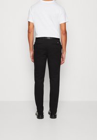 Schwarze Hosen mit schmaler Passform, aus glattem Stoff gefertigt. Verfügen über eine Gesäßtasche am Bund und sind mit schwarzen Schuhen kombiniert. Weißes T-Shirt sichtbar.