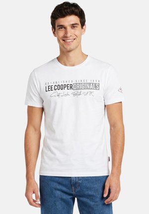 Jeune homme souriant portant un t-shirt blanc Lee Cooper Originals avec un texte noir et gris, associé à un jean bleu, debout devant un fond blanc.