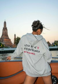 Felpa grigia con una grande stampa sul retro che recita: "Un po' di ❤️ in questo mondo brutale (ma non troppo)," indossata su una barca.