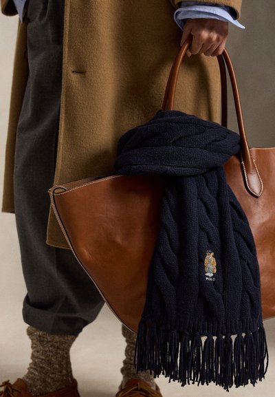 Cachecol azul-marinho de malha com padrão de trança e bordas com franjas, apresentando um logótipo bordado, pendurado numa mala tote de couro castanho.