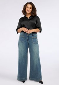 Donna sorridente che indossa una blusa nera in satin, jeans blu a gamba larga con tasche frontali e dettagli in catena, e scarpe nere a punta, in piedi.