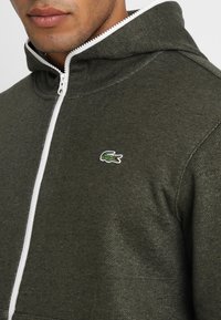 Man med mörkgrön zip-up hoodie med vit dragkedja och liten grön krokodil-logotyp på bröstet.