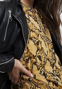 Giacca da biker nera in pelle con cerniere argentate indossata sopra un vestito con motivo a serpente giallo e marrone. Il vestito presenta un tessuto testurizzato e una chiusura con lacci.