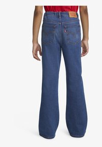 Jeans en denim avec une jambe évasée, de couleur bleu foncé, taille élastique, et deux poches arrière avec un logo brodé et une patch en cuir tan.