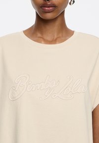 Bežinis medvilnės marškinėliai su laisvu siluetu. Priekyje yra išsiuvinėtas logotipas "Bimba y Lola" kontrastinga siūle.