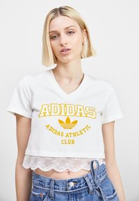 Balta trumpa marškinėliai su geltonu "ADIDAS" logotipu ir tekstu, su nėrinių apdaila apačioje ir V formos iškirpte.