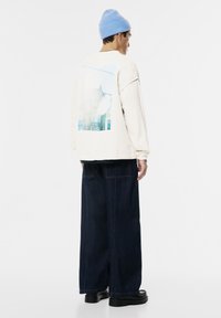 Uomo con un berretto azzurro chiaro, una maglietta a maniche lunghe crema con una stampa di un edificio sulla schiena, jeans scuri larghi e scarpe nere, in orizzontale.