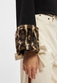 Top preto com um tecido texturizado, apresentando um punho em pelagem sintética com padrão de leopardo. O punho tem uma aparência suave e felpuda. Calças de cor clara são visíveis.