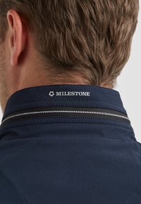 Marineblaue Jackenkragen mit weißem und schwarzem Streifendetail, auf der Rückseite mit einem gestickten Logo "MILESTONE", glatte Textur.