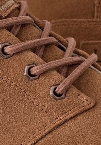 Primer plano de un zapato de ante marrón con cordones gruesos y redondos pasados por ojales metálicos hexagonales.