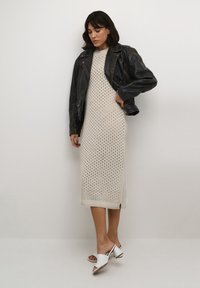 Strickkleid in Cremeton mit Spitzenmuster, kombiniert mit einer schwarzen Lederjacke. Verfügt über seitliche Schlitze und weiße offene Mules.