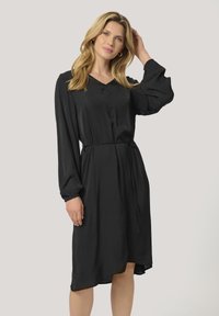 Robe noire avec un décolleté en V, de longues manches ballon et une taille ceinturée. Fabriquée en tissu lisse avec un design fluide et un ourlet asymétrique.