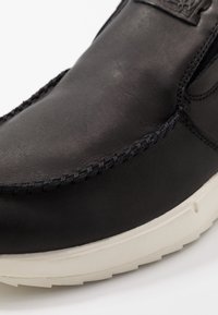 Lähikuva mustast nahast kingast, mille varvas on mustade õmblustega ja paks valge kummitald, valgel taustal.