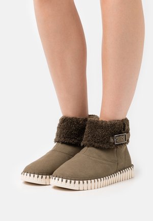 Green suede ankle boots with a fuzzy brown cuff, featuring a decorative metal buckle and a textured white sole with serrated edges.