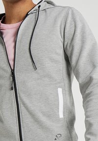 Sudadera gris con cremallera y capucha ajustable, bolsillo de acento blanco y textura suave. Presenta una cremallera negra y un logotipo en el dobladillo.