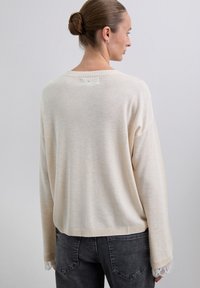 Frau mit einem Haarknoten, die einen cremefarbenen Strickpullover mit Spitzenärmeln und schwarze Jeans trägt, mit dem Rücken zu einem schlichten hellen Hintergrund.