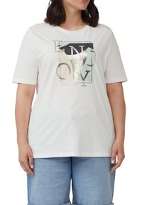 Frau trägt ein weißes Kurzarmhemd mit abstraktem geometrischen Design und Buchstaben, kombiniert mit hellblauen Jeansshorts.