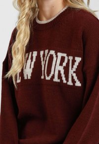 Maglione bordeaux con scritta a maglia bianca "NEW YORK". Presenta una scollatura rotonda e polsini a coste, lavorazione a maglia testurizzata. Design semplice a maniche lunghe.