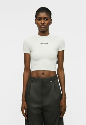 Γυναίκα που φοράει λευκό κοντό t-shirt με την επιγραφή «Palm Angels» και μαύρο παντελόνι με ψηλή μέση, στέκεται μπροστά σε απλό λευκό φόντο.