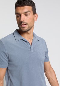 Polo shirt van zacht blauw fabric, met een klassieke kraag, korte mouwen en een ontspannen pasvorm. De oppervlakte textuur lijkt iets fuzzy.