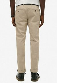 Pantalones de algodón beige con corte recto, que cuentan con dos bolsillos traseros y dobladillos enrollados. Combinados con zapatos negros, que muestran una textura sutil.