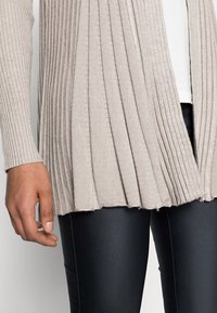 Cardigan beige clair avec texture côtelée et plis verticaux, devant ouvert et ourlet brut. Porté sur un legging noir, mettant en avant un look décontracté.