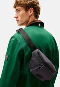 Bolso bandolera gris con acabado mate, que cuenta con un compartimento principal con cierre, un diseño elegante y un bolsillo lateral más pequeño, llevado estilo cruzado.