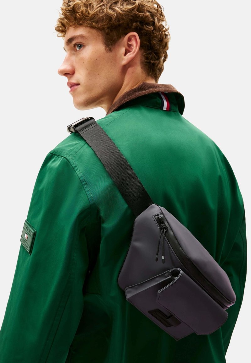 Bolso bandolera gris con acabado mate, que cuenta con un compartimento principal con cierre, un diseño elegante y un bolsillo lateral más pequeño, llevado estilo cruzado.