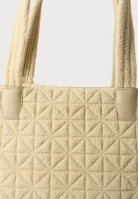 Sac tote matelassé beige en tissu texturé, présentant des motifs triangulaires et des détails en cuir aux poignées.
