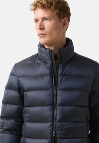 Mladý muž s kudrnatými vlasy, oblečený v zapínané námořnické modré péřové bundě, dívající se do strany na jednobarevném pozadí.