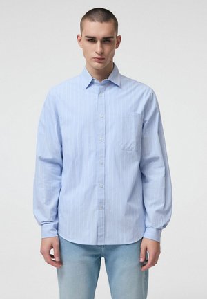 Młody mężczyzna ubrany w jasnoniebieską, zapinaną na guziki koszulę z długim rękawem i białe pionowe paski oraz jasnoniebieskie dżinsy, stojący na jednolitym tle.