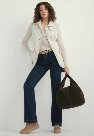 Kobieta ubrana w beżową pikowaną kamizelkę, kremowy prążkowany sweter, ciemnoniebieskie dżinsy, beżowe tenisówki, trzymając po bokach dużą ciemnobrązową torebkę.