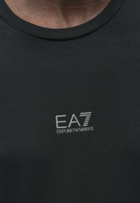 Fekete pamut póló környakú kivitelben. Bal mellrészen szürke, "EA7 EMPORIO ARMANI" feliratú nyomtatott logóval. Sima tapintású anyag.