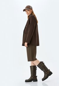 Blazer marrone oversize con texture semplice, abbinato a una gonna a quadri marrone al ginocchio. Alti stivali di pelle marrone con fibbie.