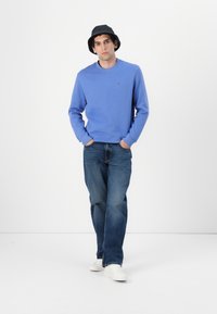 Felpa in cotone azzurro chiaro, con collo a giro, abbinata a jeans in denim blu scuro e sneakers bianche. Cappello a secchiello nero completa l'outfit.