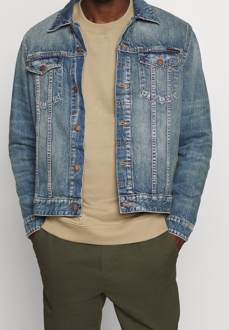 Jeansjacka i ljusblått med en urtvättad look, med bruna knappar och två bröstfickor. Bärs över en beige crewneck-tröja.