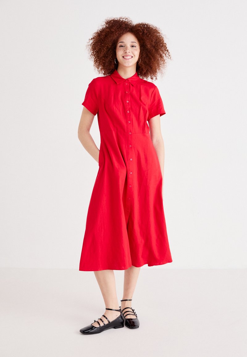 Κόκκι�νο φόρεμα κοντομάνικο με κουμπιά μπροστά και τσέπες. Ροή σε σχήμα Α, φτιαγμένο από υφασμάτινη δομή, φορεμένο με μαύρες πλατφόρμες.