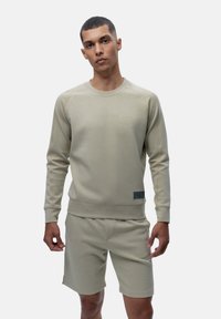 Light beige sweatshirt with ribbed neckline and cuffs, featuring subtle branding and a textured logo patch, paired with matching shorts.
