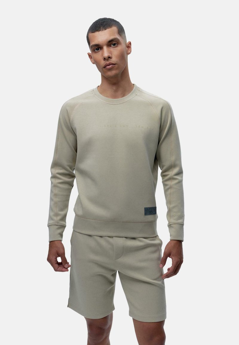 Light beige sweatshirt with ribbed neckline and cuffs, featuring subtle branding and a textured logo patch, paired with matching shorts.