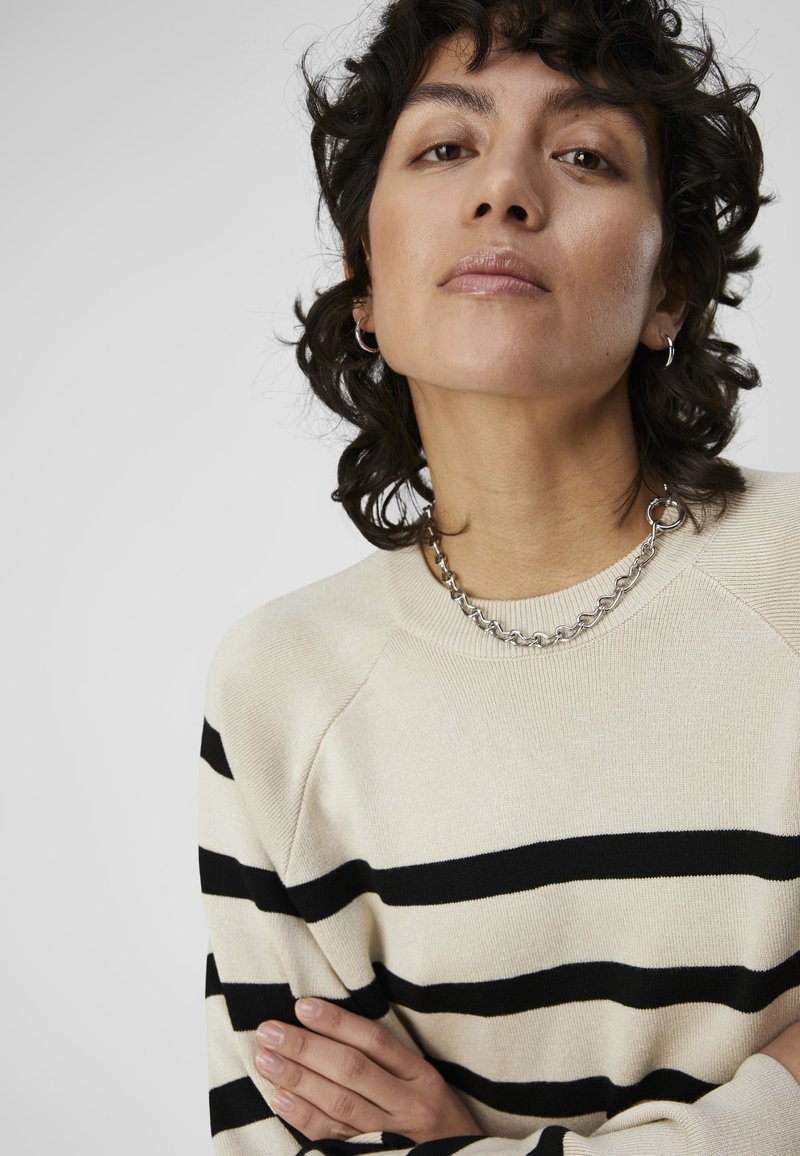 Beige Pullover mit schwarzen Streifen, mit Rundhalsausschnitt und geripptem Design. Das Model trägt eine silberne Kettenhalskette und hat kurzes, lockiges Haar.