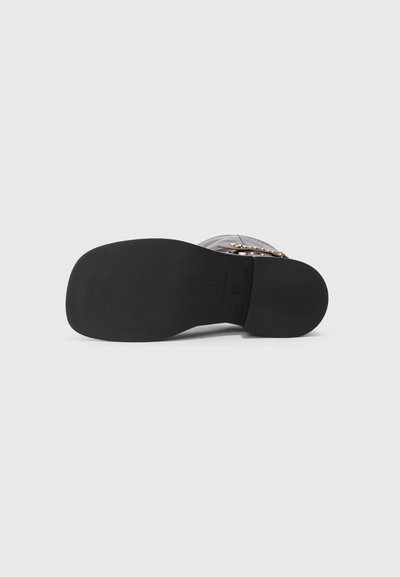 Semelle en caoutchouc noir d'une chaussure, avec une texture lisse et un léger motif de crantage. Matériau supérieur transparent avec un accent de boucle décorative.