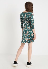 Blumenkleid in Grün mit Dreiviertelärmeln, das eine taillierte Passform und Raffung an der Taille aufweist. Kombiniert mit schwarzen Sneakers.