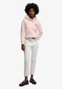 Różowa bluza z kapturem z kieszenią kangurką i zielonym logo, noszona na niebiesko-białej pasiastce, zestawiona z białymi dżinsami i ciemnymi mokasynami.