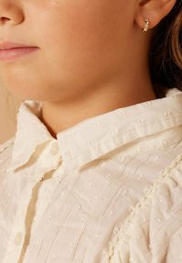 Gouden studs met een klein ananasontwerp en juwelenaccenten, gedragen met een witte shirt met textuur en een knoopkraag.