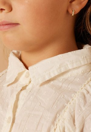 Boucles d'oreilles en or avec un petit design d'ananas et des accents de bijoux, portées avec une chemise blanche texturée à col boutonné.