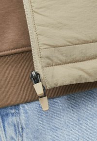 Tissu de doudoune beige avec texture matelassée, comportant une fermeture éclair beige clair et des accessoires noirs, accompagné d'un poignet côtelé marron et d'un denim bleu clair.